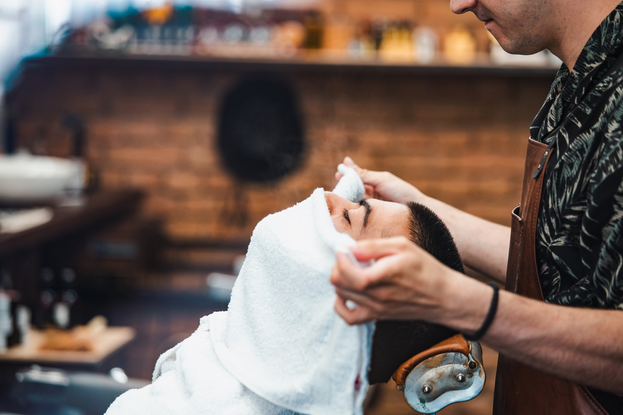 Barber covers the face of a man with a hot towel. Traditional ritual of shaving the beard with hot and cold compresses in a old style barber shop. Client with hot towel on face before shaving in barber shop